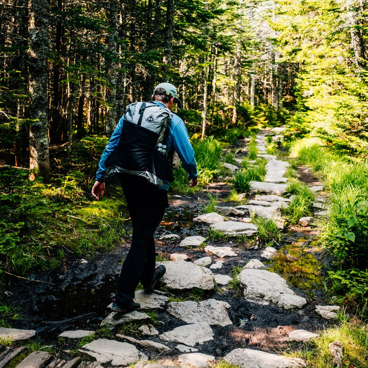 A hiker wearing a black and white backpack walks along a rocky forest trail surrounded by green trees and foliage.