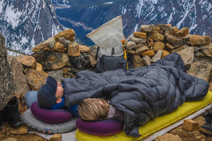 Two people camping on a mountain ridge, lying under a dark gray sleeping bag on a yellow sleeping pad with purple pillows. A white tent, backpack, and stone wall are visible in the background with a snowy mountain landscape.