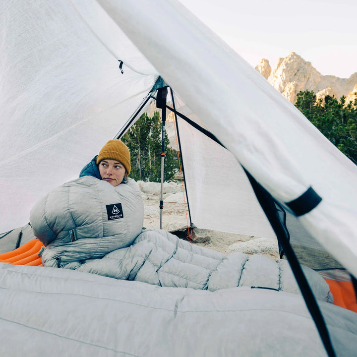 Person lying in a sleeping bag inside a tent with mountains in the background