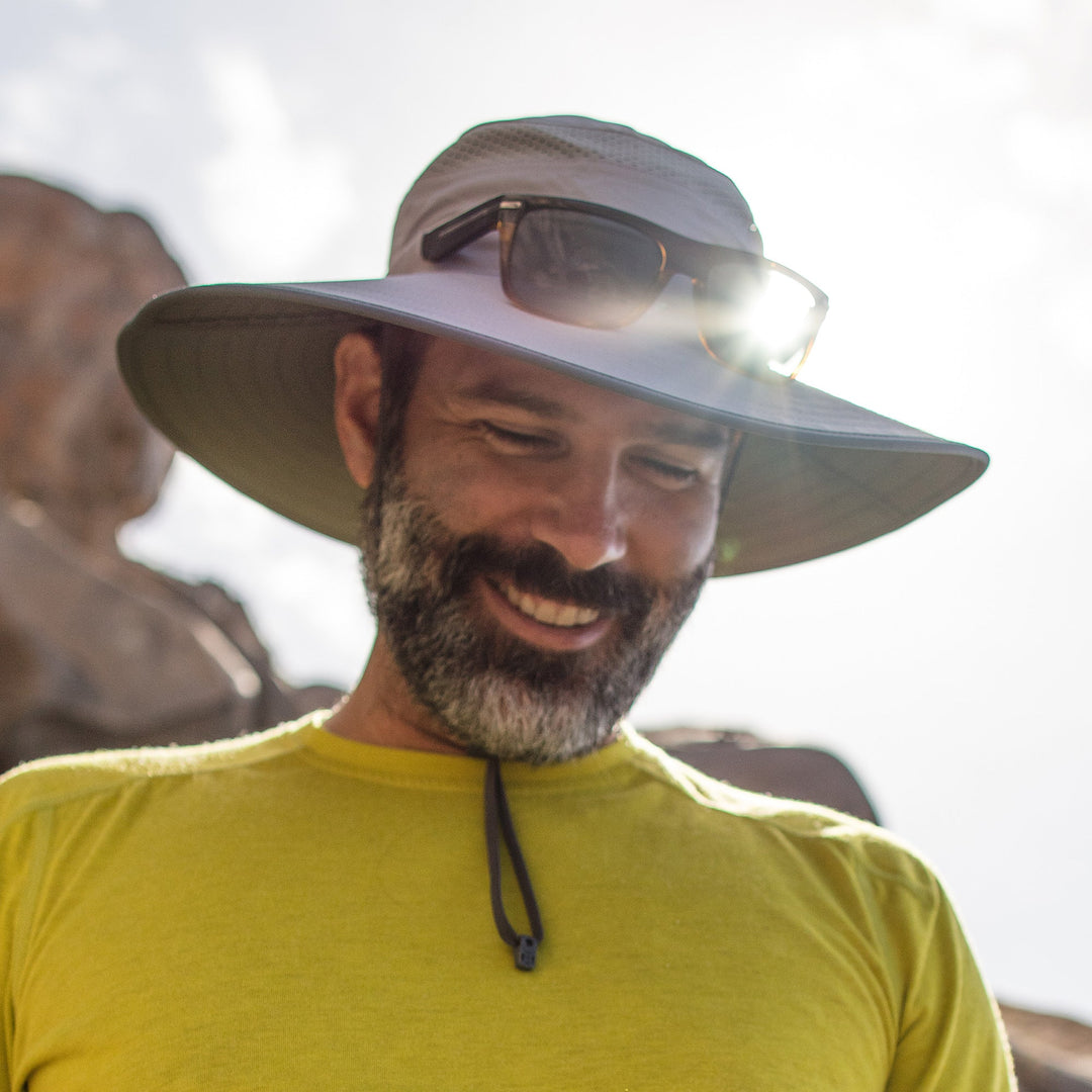 Man wearing a wide-brimmed hat with sunglasses on top, smiling outdoors.