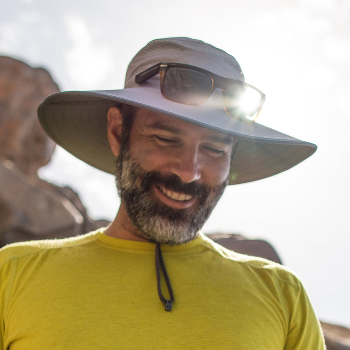 Man wearing a wide-brimmed hat with sunglasses on top, smiling outdoors.
