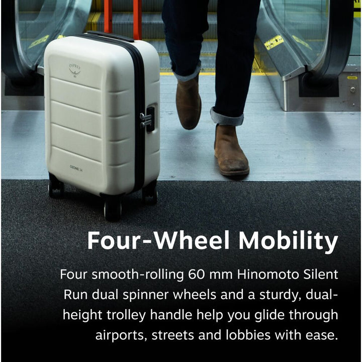 A cream-colored hardshell suitcase with black trim and four wheels stands on a dark floor in front of an escalator. A person&