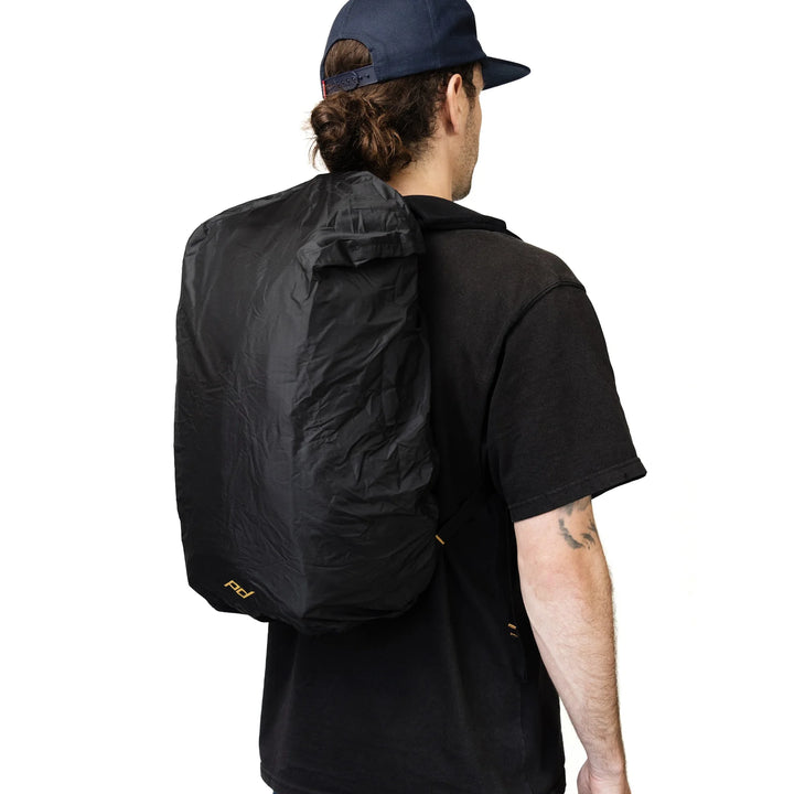 A man shown from behind wearing a black backpack with a matching black rain cover, a black t-shirt, and a navy blue baseball cap, against a solid white background.