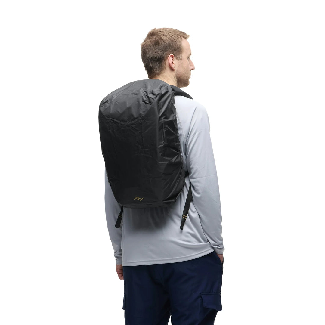 A man shown from the back wearing a black waterproof backpack cover over a black backpack, dressed in a light gray long-sleeve shirt and navy pants, against a solid white background.
