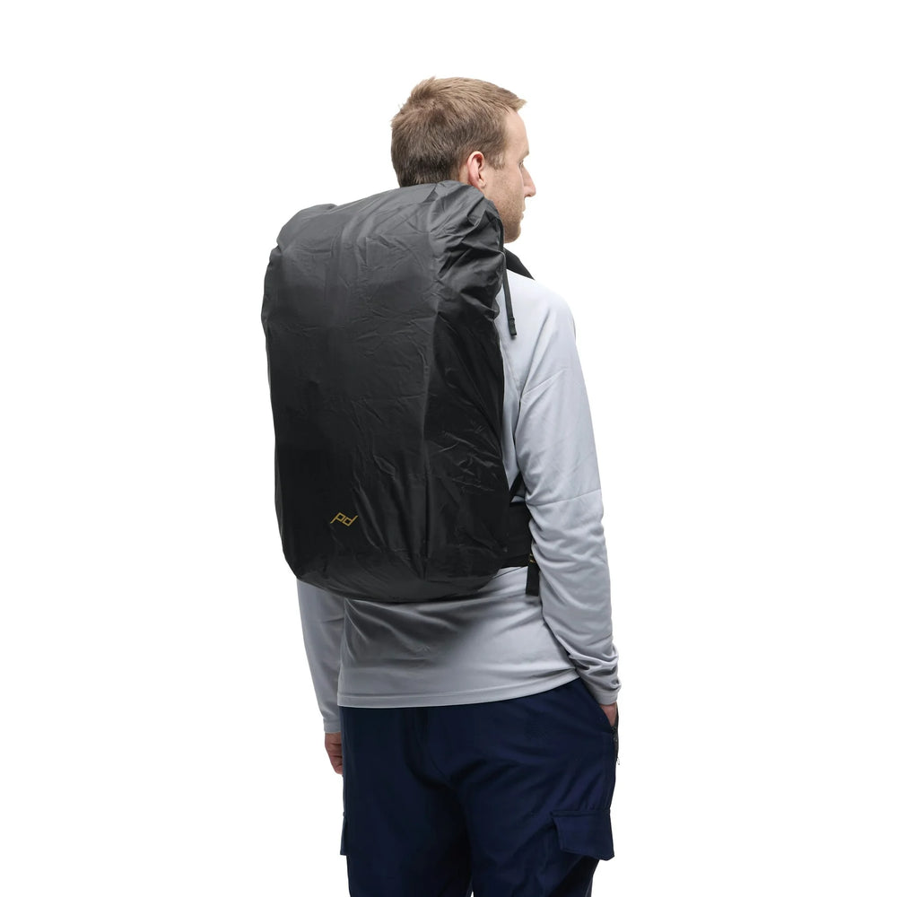 A man shown from the back wearing a black backpack rain cover over a gray long-sleeve shirt and navy pants, against a plain white background.