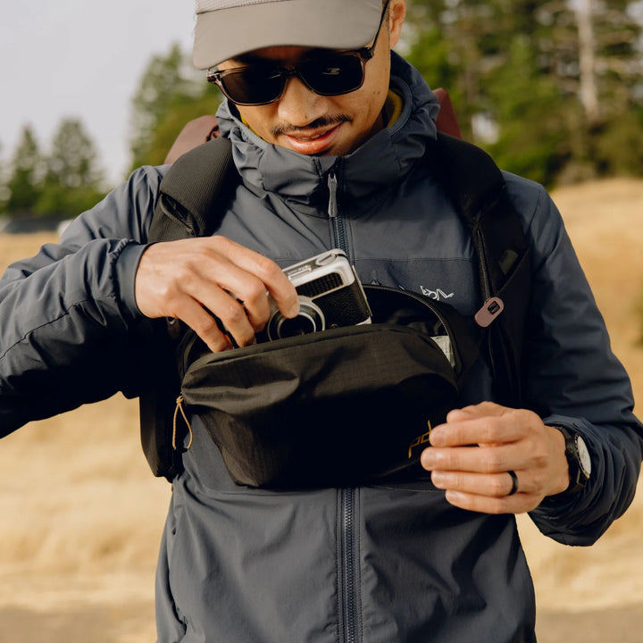 Person in outdoor setting wearing a jacket and sunglasses, holding a camera and a bum bag.