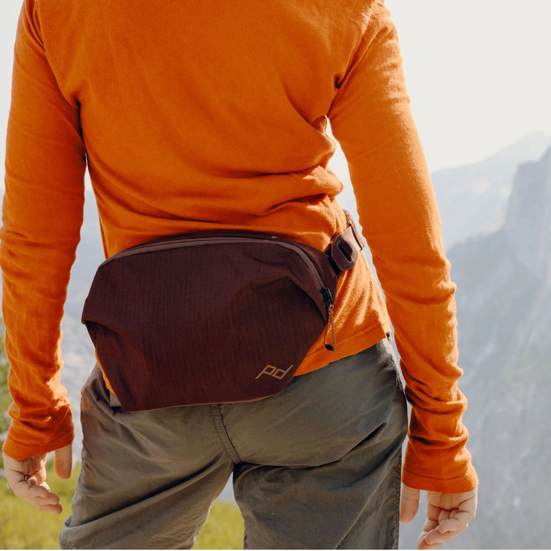 Person wearing an orange long-sleeve shirt and brown waist bag with mountains in the background