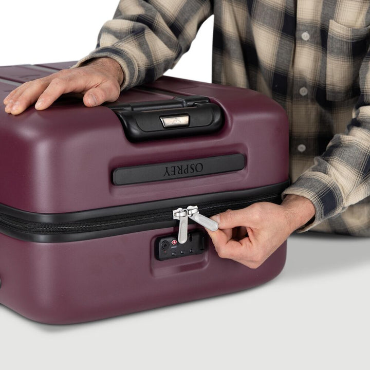 A person wearing a beige and black plaid flannel shirt is shown from the chest down, interacting with a burgundy Osprey hard-shell suitcase. Their left hand rests on the suitcase top, and their right hand is turning the silver zipper pull.