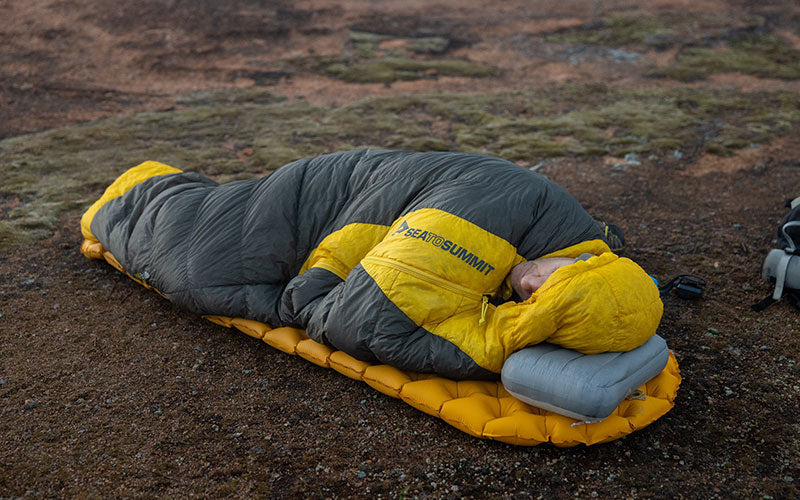 Person in a sleeping bag with their head on a grey inflatable pillow in the outdoors