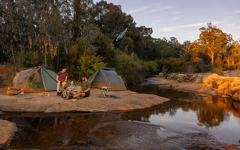 Two green and brown tents pitched on the bank of a creek, with some three people sitting in front of them.