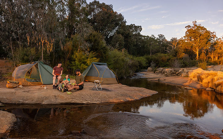 Two green and brown tents pitched on the bank of a creek, with some three people sitting in front of them.