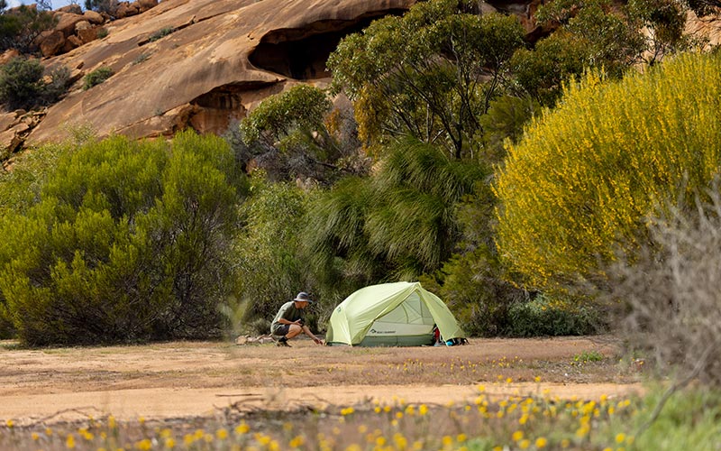 Man crouching next to a green tent in a outdoors setting with trees and rocks.