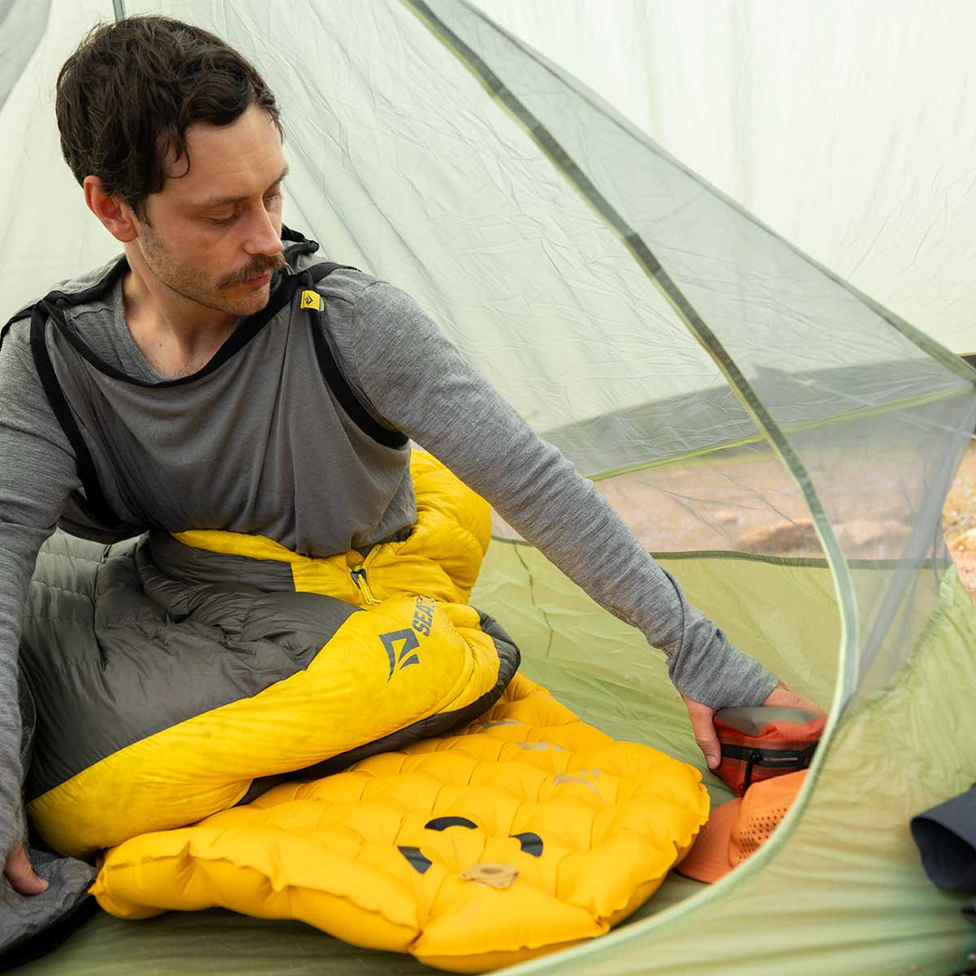 Person sitting inside a tent with yellow sleeping bag and camping gear.