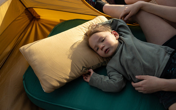 Child sprawled on a green sleeping mat with head on a brown pillow in a yellow tent