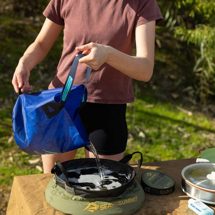 Person pouring water from a blue bag into a pot on a camping stove outdoors.