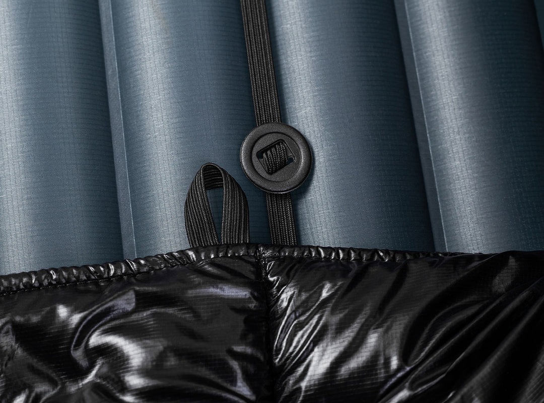 A black quilted puffer sleeping quilt shown against a navy blue sleeping pad  with an elastic strap and button 