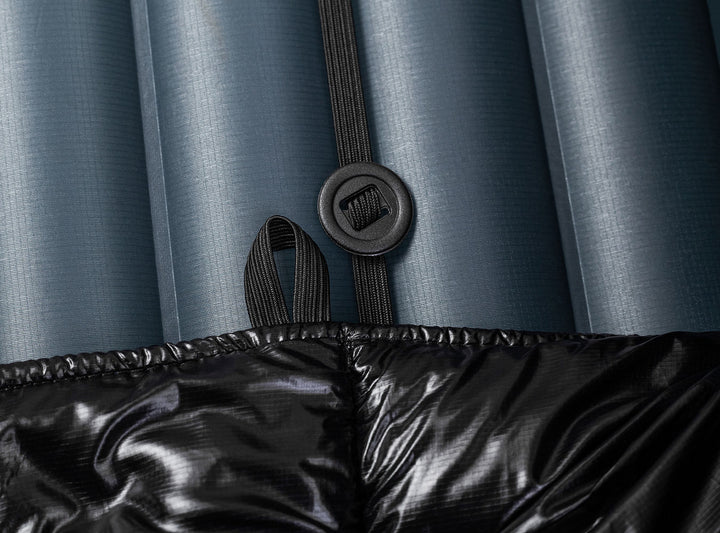 A black quilted puffer sleeping quilt shown against a navy blue sleeping pad  with an elastic strap and button 