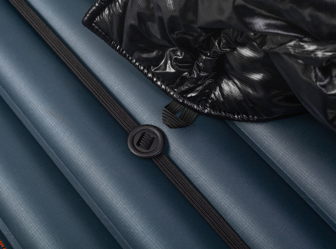 A black quilted puffer sleeping quilt shown against a navy blue sleeping pad  with an elastic strap and button 