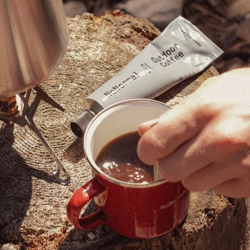 Person pouring coffee from a tube labeled 'Outdoor Coffee' into a red mug on a natural surface.