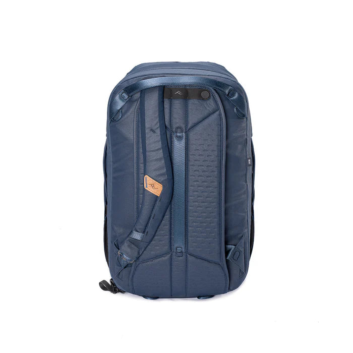 A dark blue technical backpack shown from the back, featuring padded shoulder straps, a textured back panel, and a small tan leather brand patch, set against a plain white background.