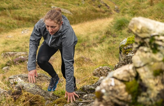 Person scrambling up a rocky hill side wearing a grey waterproof jacket